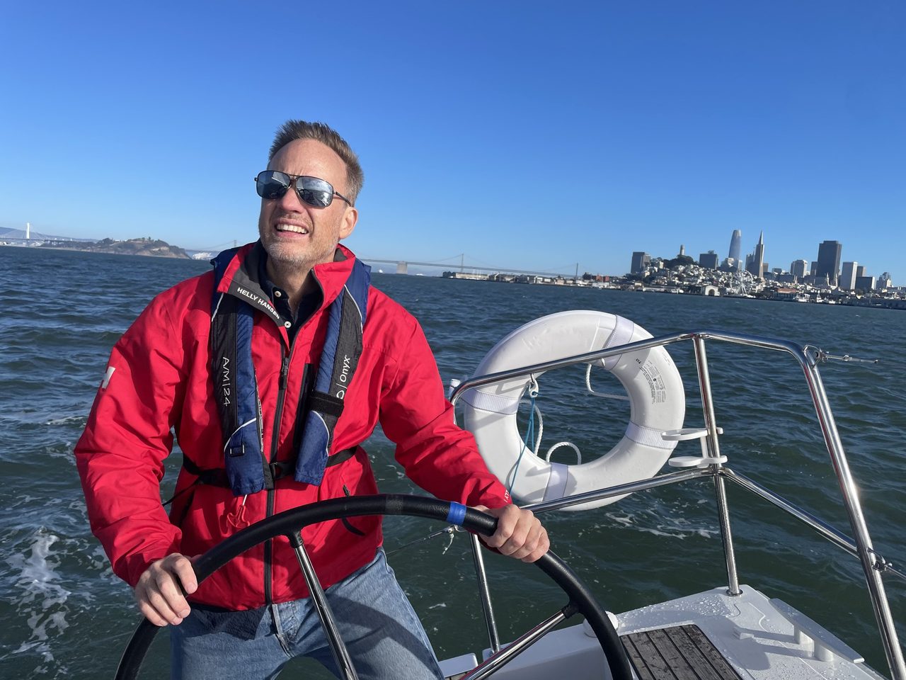 Jonathan Weedon at the helm of a sailboat on San Francisco Bay, with the city skyline and the Bay Bridge behind him